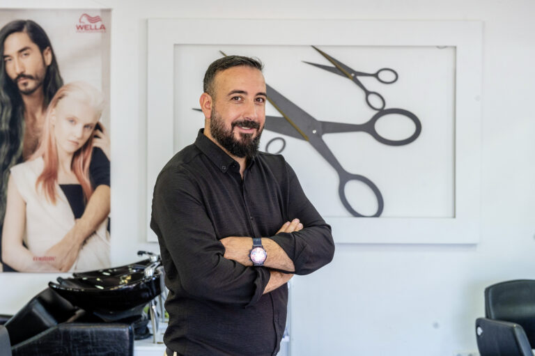 Porträt von Hasret und Abdullah Sari in ihrem Friseursalon vor einer Wand mit Scheren-Grafik.
