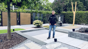 Sebastian Müller steht in seinem Mustergarten in Bargstedt, umgeben von verschiedenen Gartenmaterialien und Pflanzen.