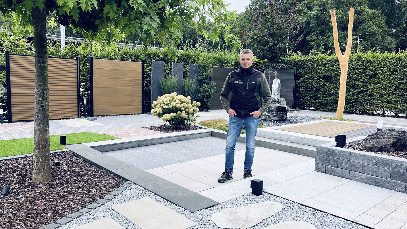 Sebastian Müller steht in seinem Mustergarten in Bargstedt, umgeben von verschiedenen Gartenmaterialien und Pflanzen.
