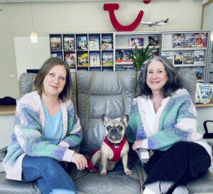 Tanja Strobel und eine Kollegin sitzen mit einem kleinen Hund auf einem Sofa in einem Reisebüro vor einem Regal mit Katalogen.
