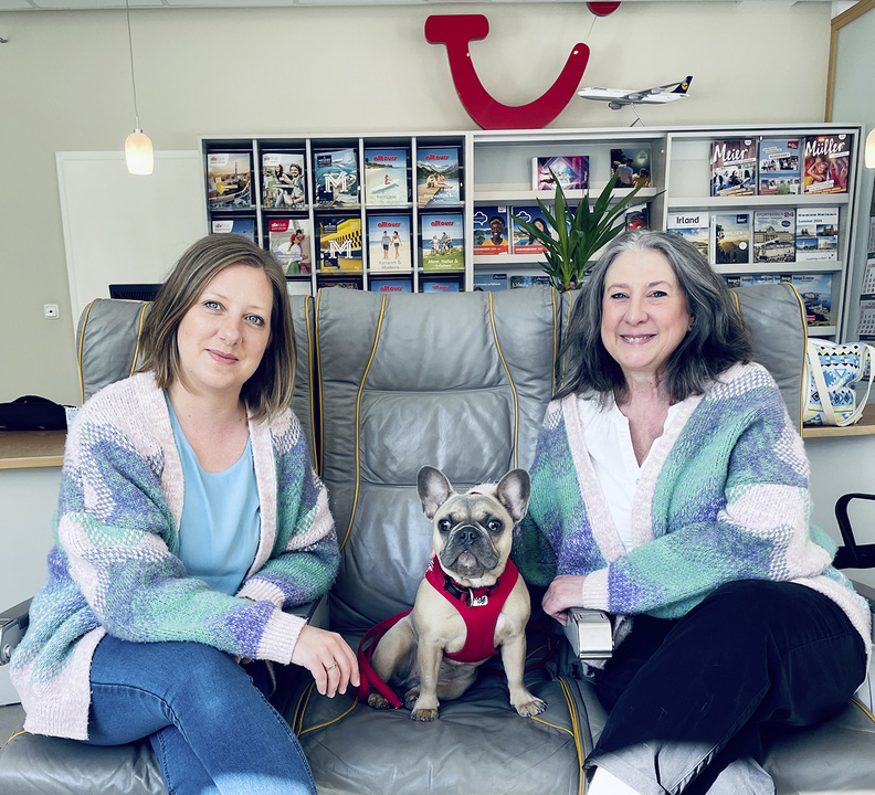 Tanja Strobel und eine Kollegin sitzen mit einem kleinen Hund auf einem Sofa in einem Reisebüro vor einem Regal mit Katalogen.