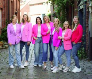 Sieben Frauen in pinken und roten Blazern stehen auf einem gepflasterten Weg zwischen Backsteingebäuden und lächeln in die Kamera