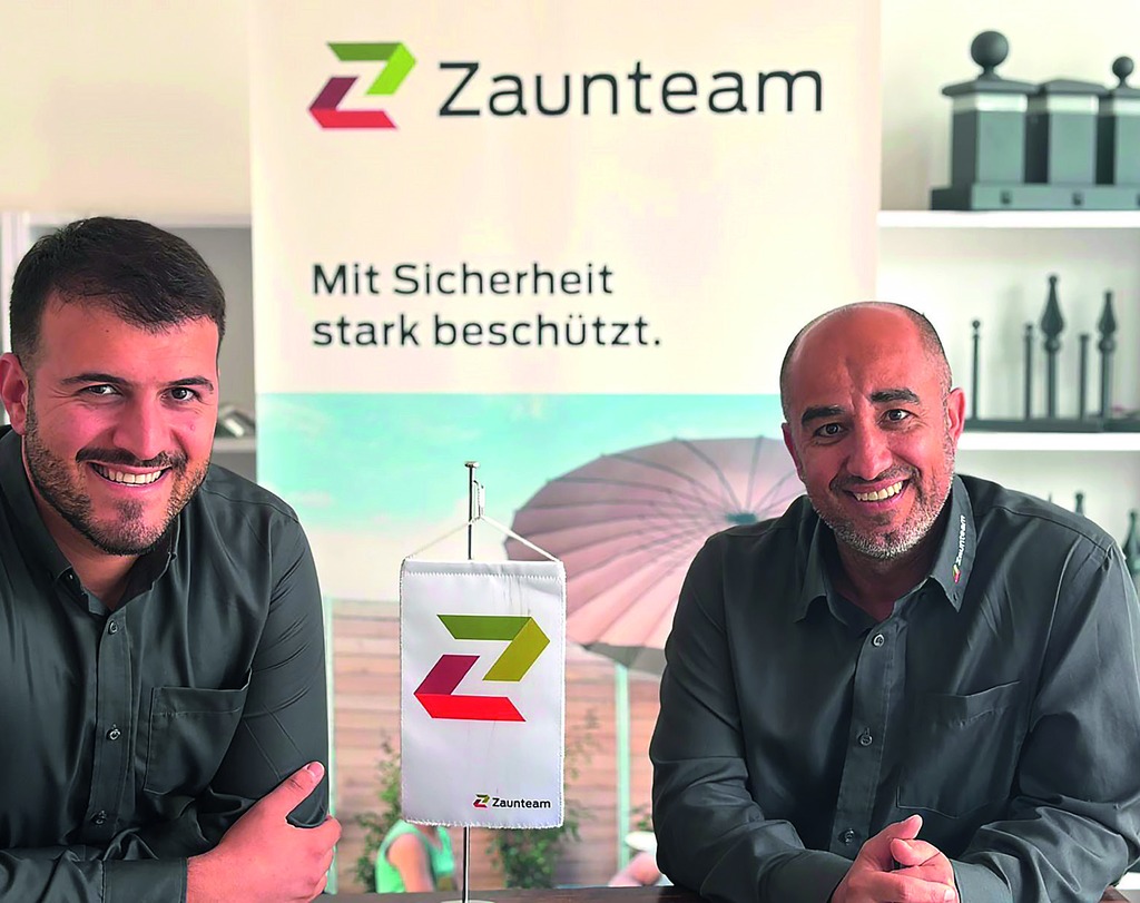 Zwei lächelnde Männer sitzen an einem Tisch vor einem Zaunteam-Banner; auf dem Tisch steht ein kleines Zaunteam-Wimpel, im Hintergrund Regale mit Zaunelementen.